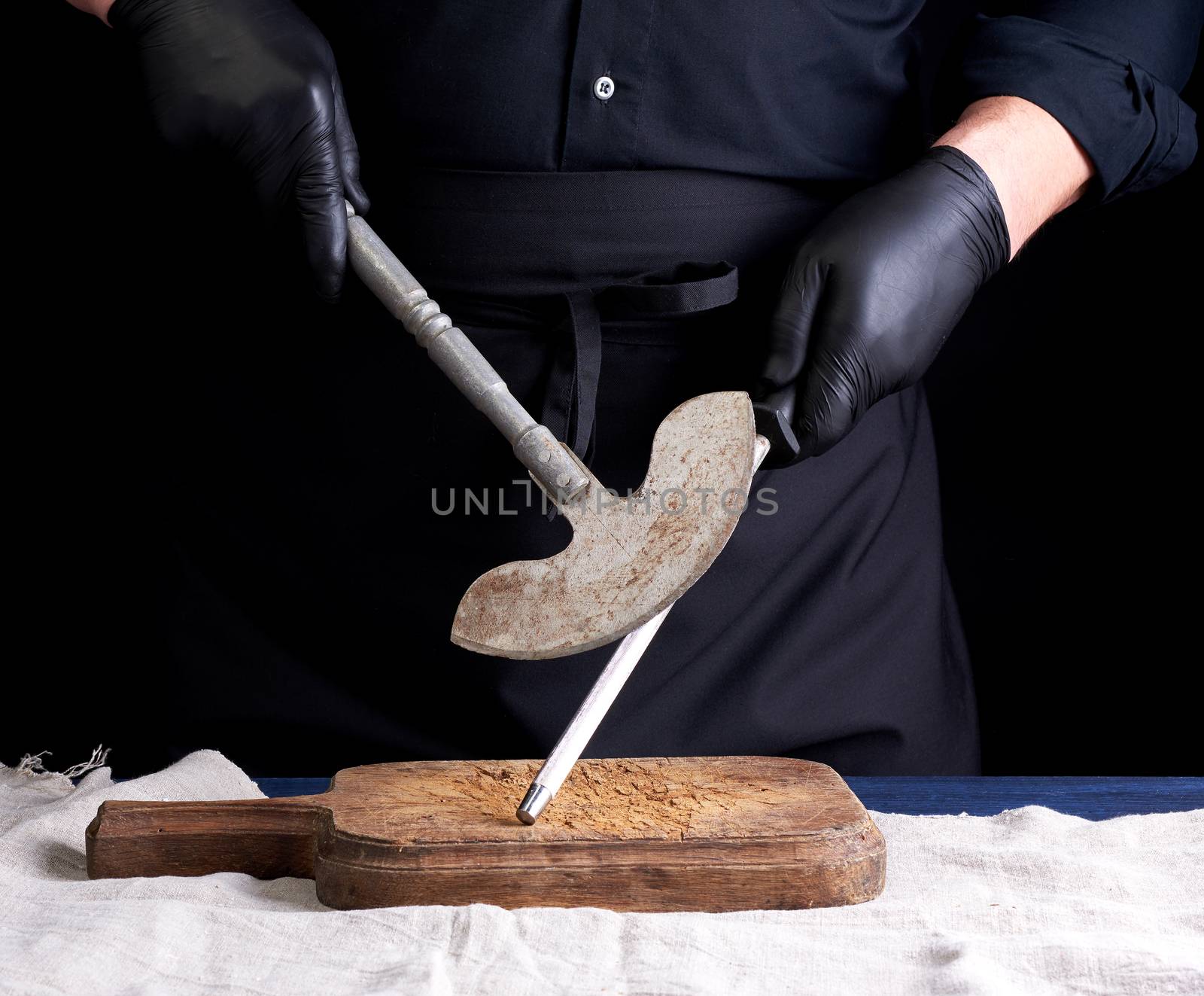 male cook in black uniform and black latex gloves holds a large sharp vintage knife for meat and vegetables and a metal sharpener for knives, black background