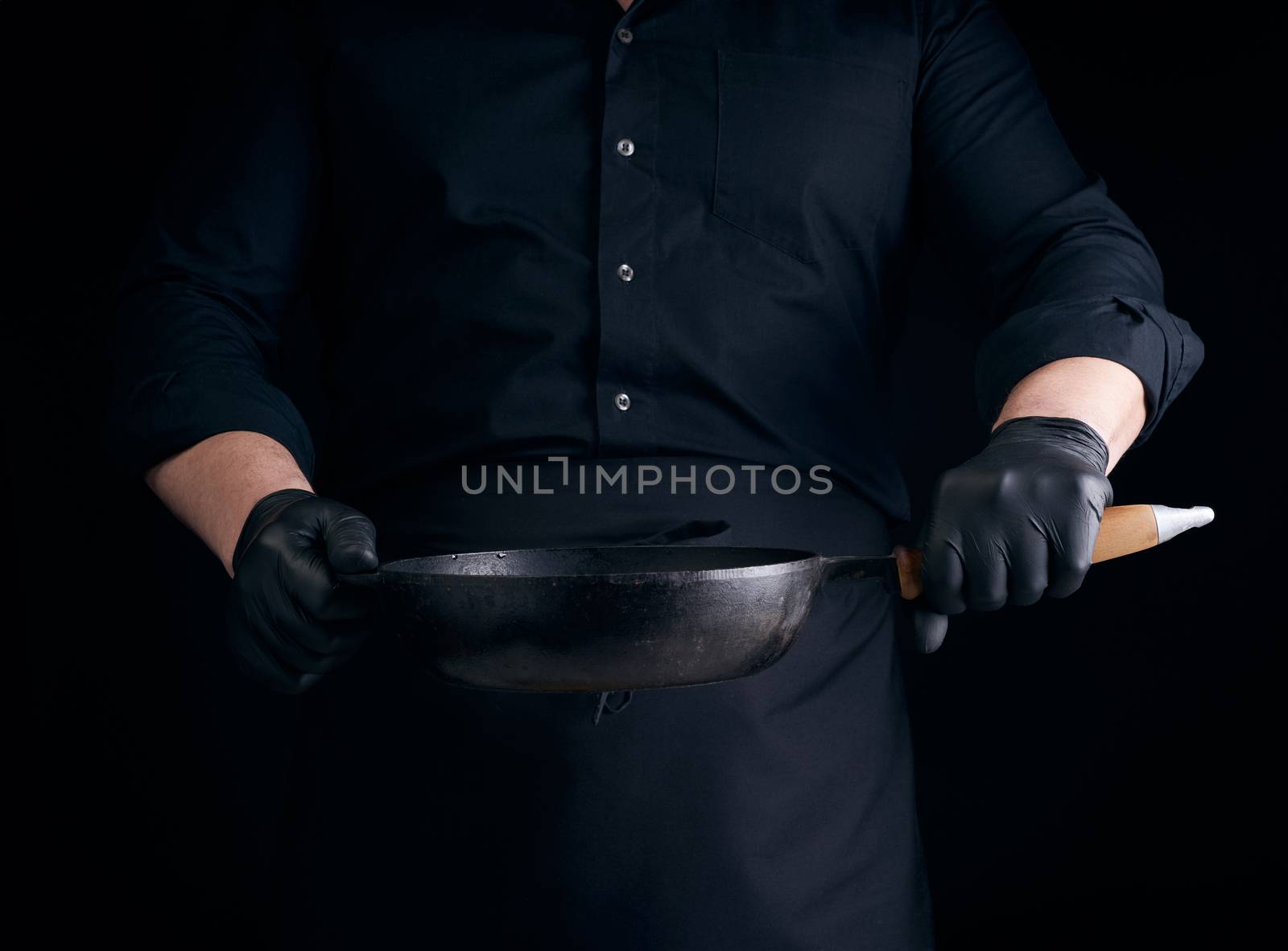 male cook in black uniform and latex gloves holds an empty round vintage black cast iron pan in front of him, low key