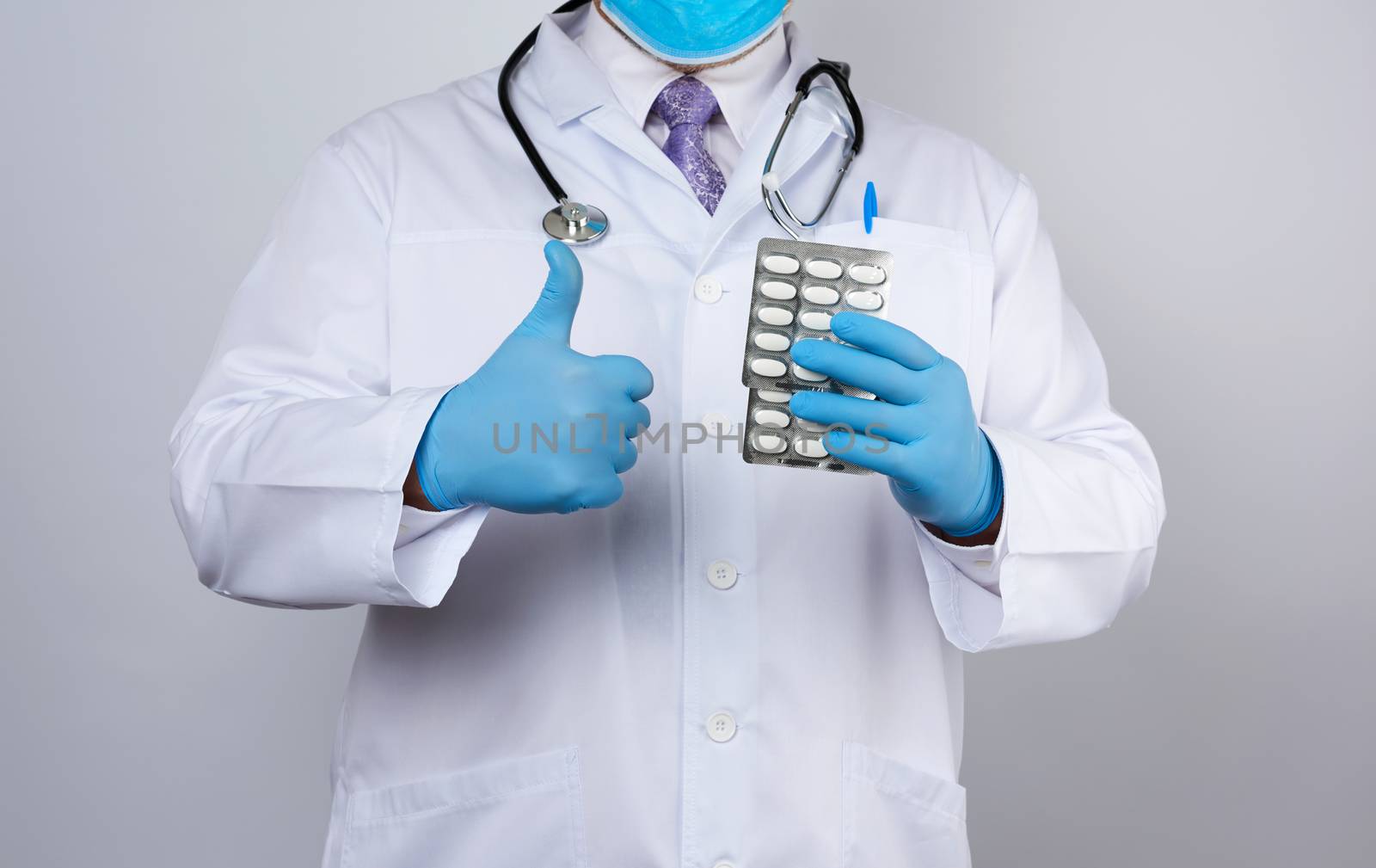 doctor therapist is dressed in a white uniform coat and blue sterile gloves is standing and holding a stack of pills in blister packs, white background
