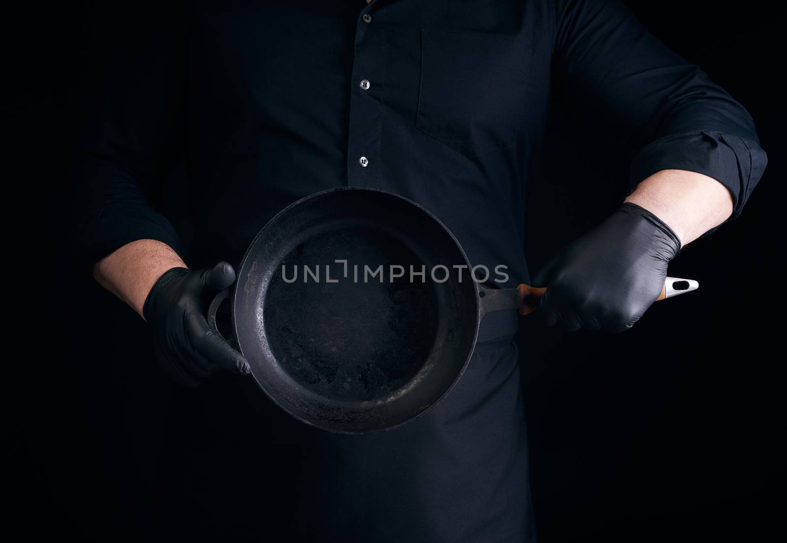male cook in black uniform and latex gloves holds an empty round vintage black cast iron pan in front of him, low key