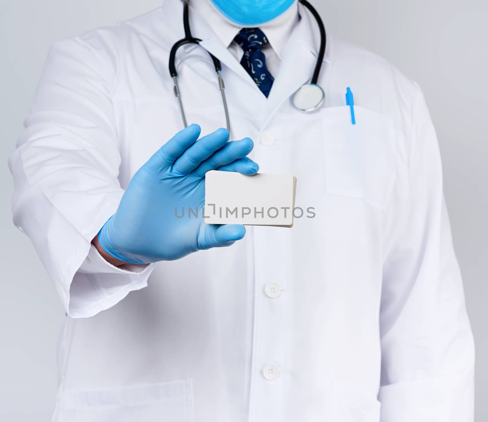 adult doctor therapist is dressed in a white robe uniform and blue sterile gloves is standing and holding a stack of empty white paper business cards, place for inscription