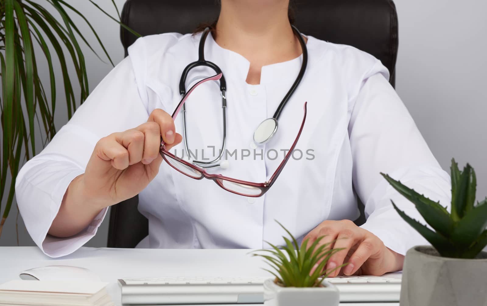 woman doctor in a white uniform, disposable medical mask sits in a chair at the table and works, holds glasses in his hands, office