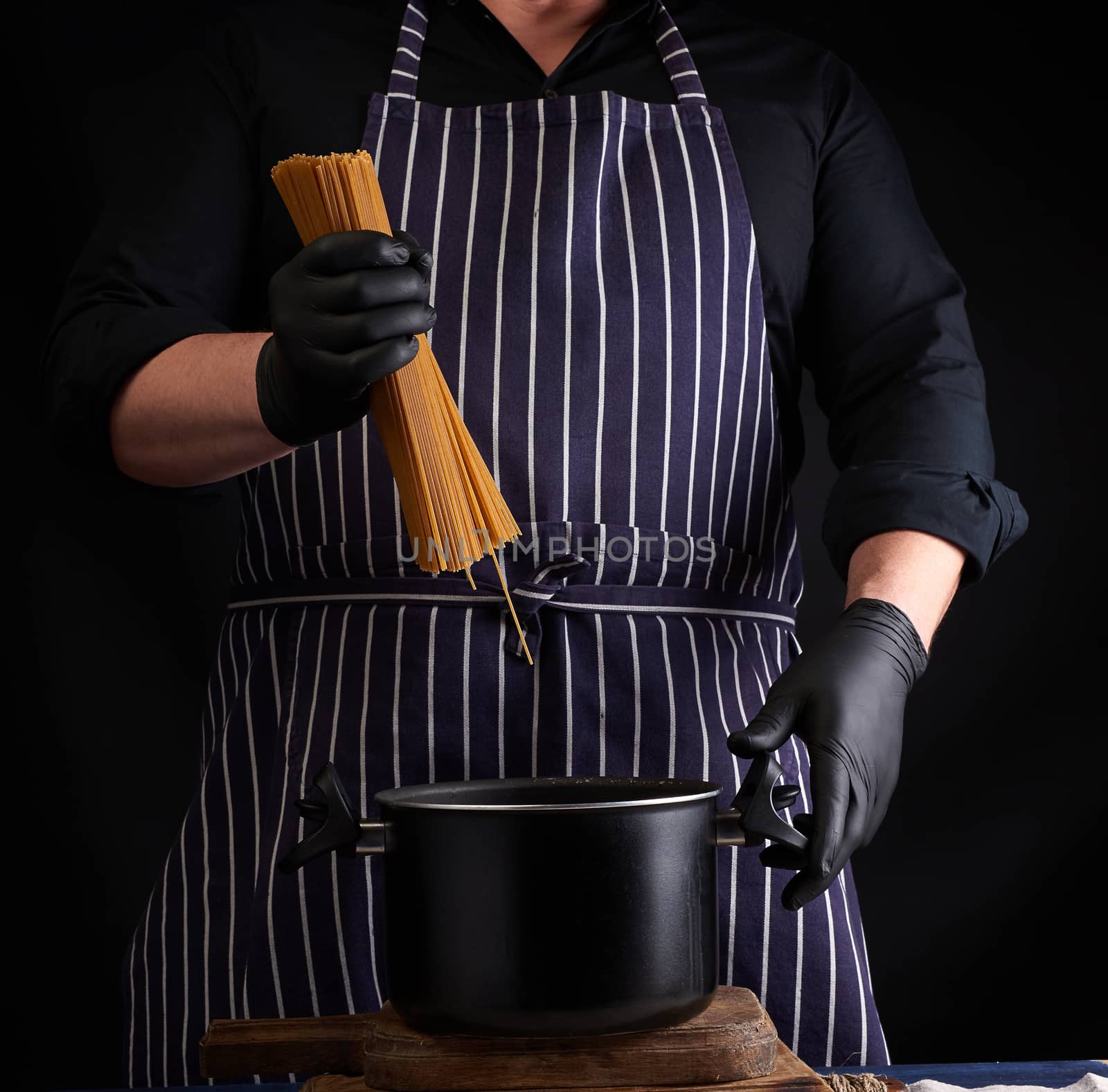 chef in black latex gloves, striped apron holds raw long spaghetti over the pan, cooking process