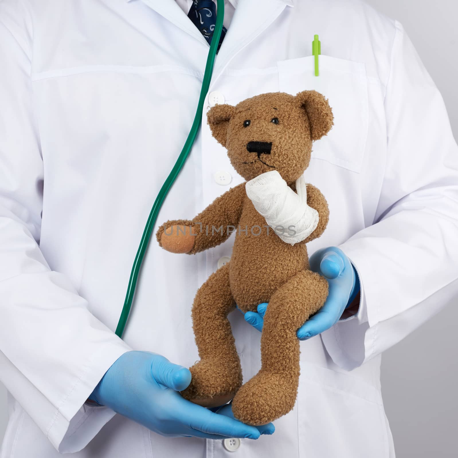 pediatrician in a white coat, blue latex gloves holds a brown teddy bear with a bandaged paw, toy treatment