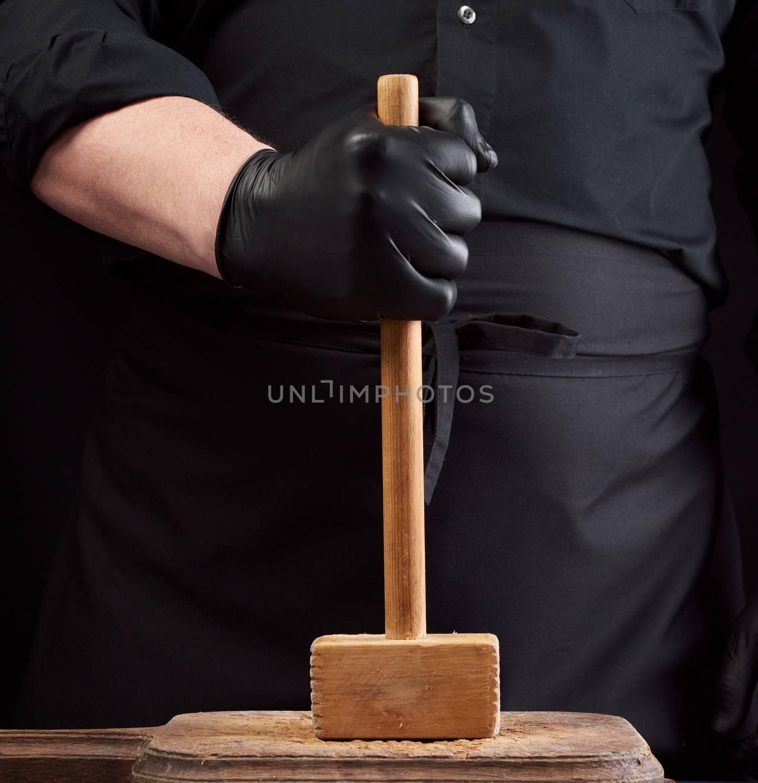 cook in black uniform holds a wooden hammer for beating meat on a vintage cutting board, black background