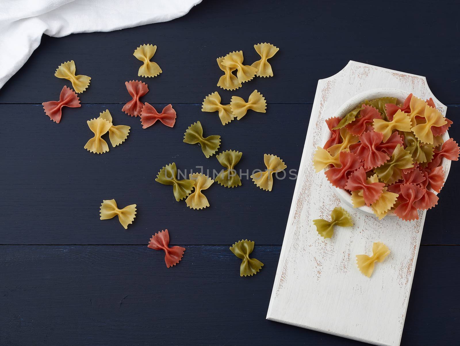 raw pasta in the form of bows in a wooden round plate and white textile napkin on a blue wooden background, top view. Vegetarian nutrition. Italian cuisine