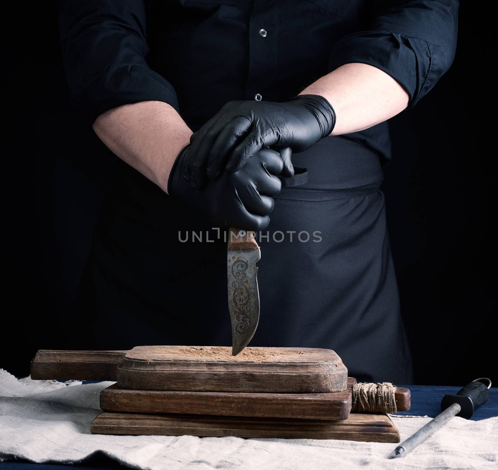 male cook in black uniform and black latex gloves holds a vintage sharp knife over a cutting board, black background