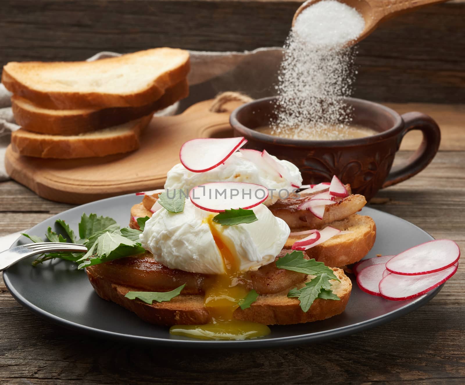 two sandwiches on a toasted white slice of bread with poached eggs, green leaves of arugula and radish in a round plate, behind a cup of coffee, morning breakfast