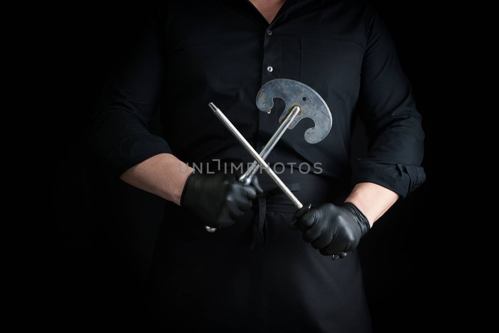 male cook in black uniform and black latex gloves holds a large sharp vintage knife for meat and vegetables and a metal sharpener for knives, black background