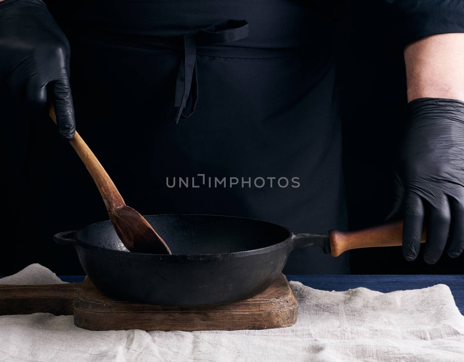 cook in black uniform and latex gloves stirs a vintage wooden spoon food in a cast-iron pan with a handle, dark background