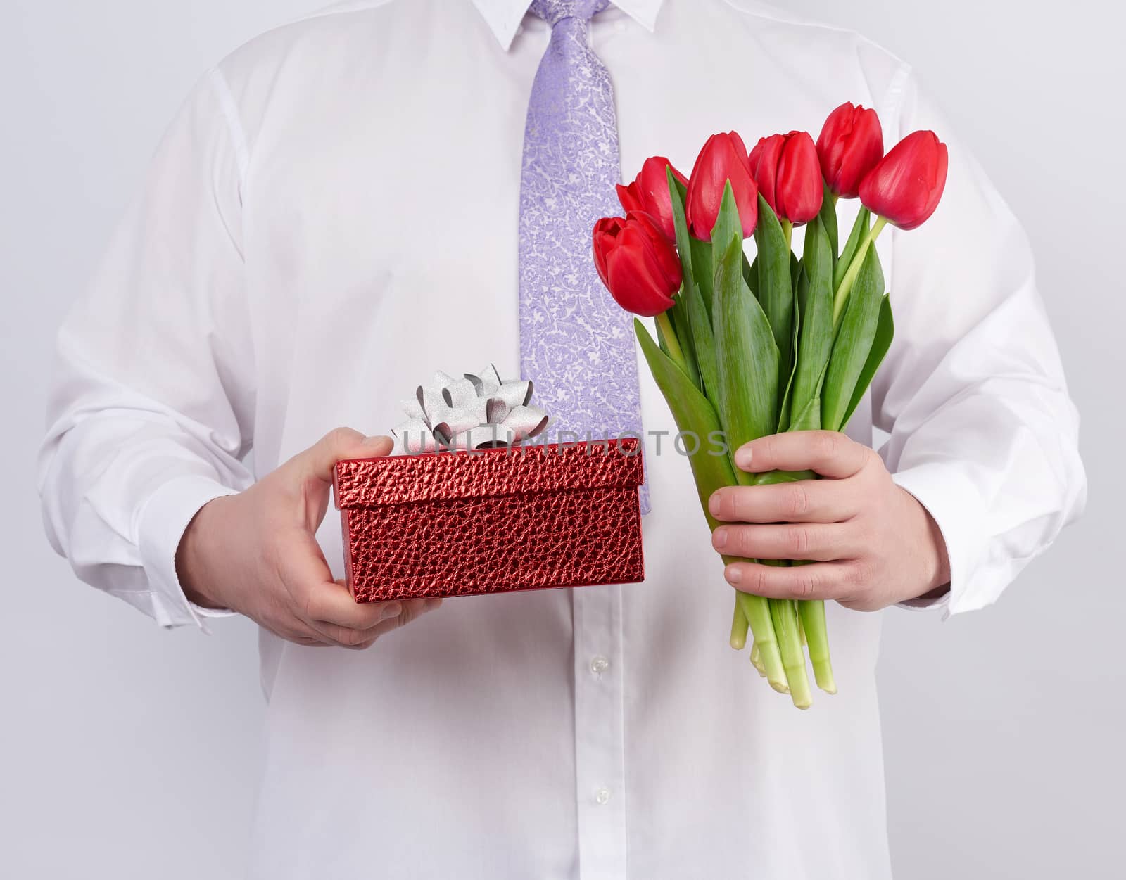 adult man in a white shirt and a lilac tie holding a bouquet of red tulips with green leaves and gift box on a white background, concept for happy birthday, anniversary, valentines day