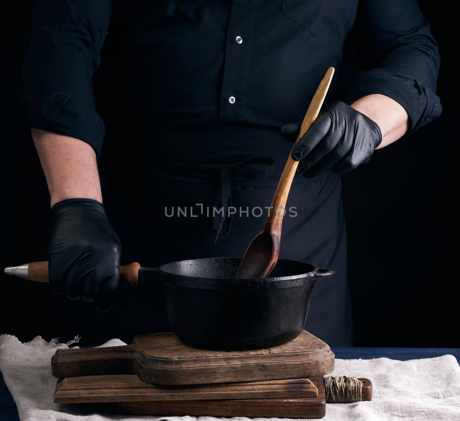 male cook in black uniform and latex gloves stirs a vintage wooden spoon food in a cast-iron pan with a handle, dark background