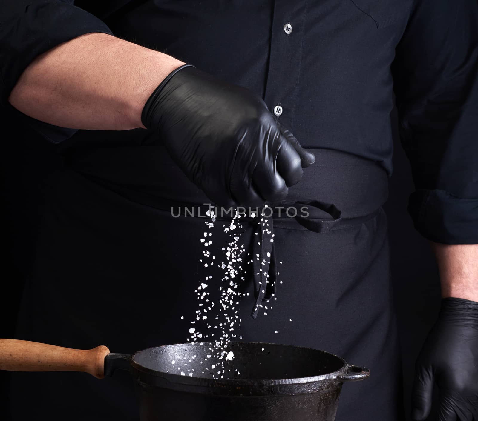 male cook in black uniform and latex gloves salt food in a black cast-iron pan, salt crystals froze in the air, low key
