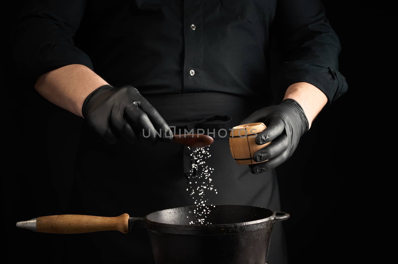 male cook in black uniform and latex gloves salt food in a black cast-iron pan, salt crystals froze in the air, low key