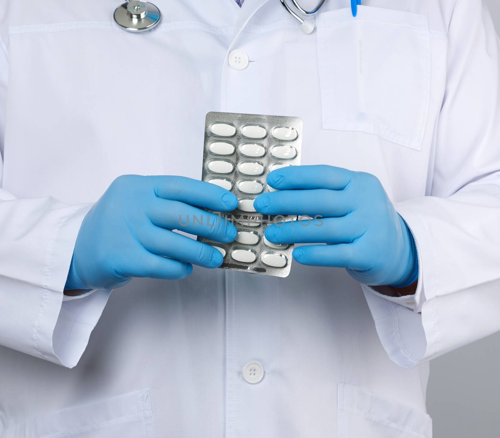 doctor therapist is dressed in a white uniform coat and blue sterile gloves is standing and holding a stack of pills in blister packs, white background