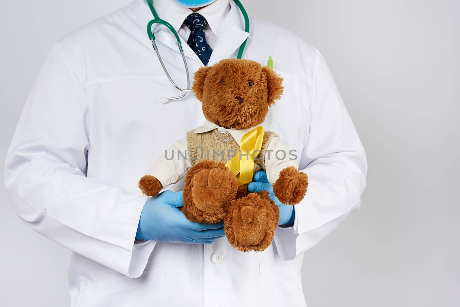 pediatrician in white coat, blue latex gloves holds a brown teddy bear with a yellow ribbon on a sweater, concept of the fight against childhood cancer. problem of suicides