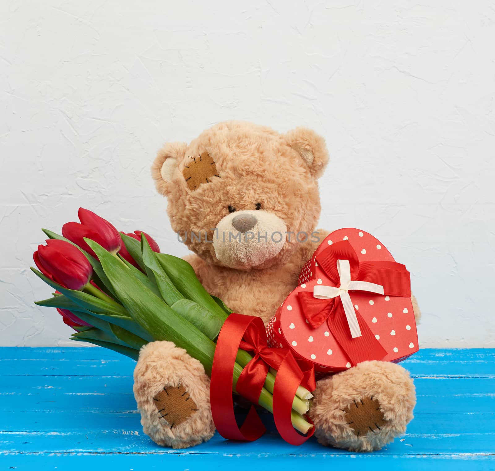 cute brown teddy bear sits on a blue wooden background, bouquet of red tulips, red box, festive backdrop for birthday, Valentine's day, anniversary