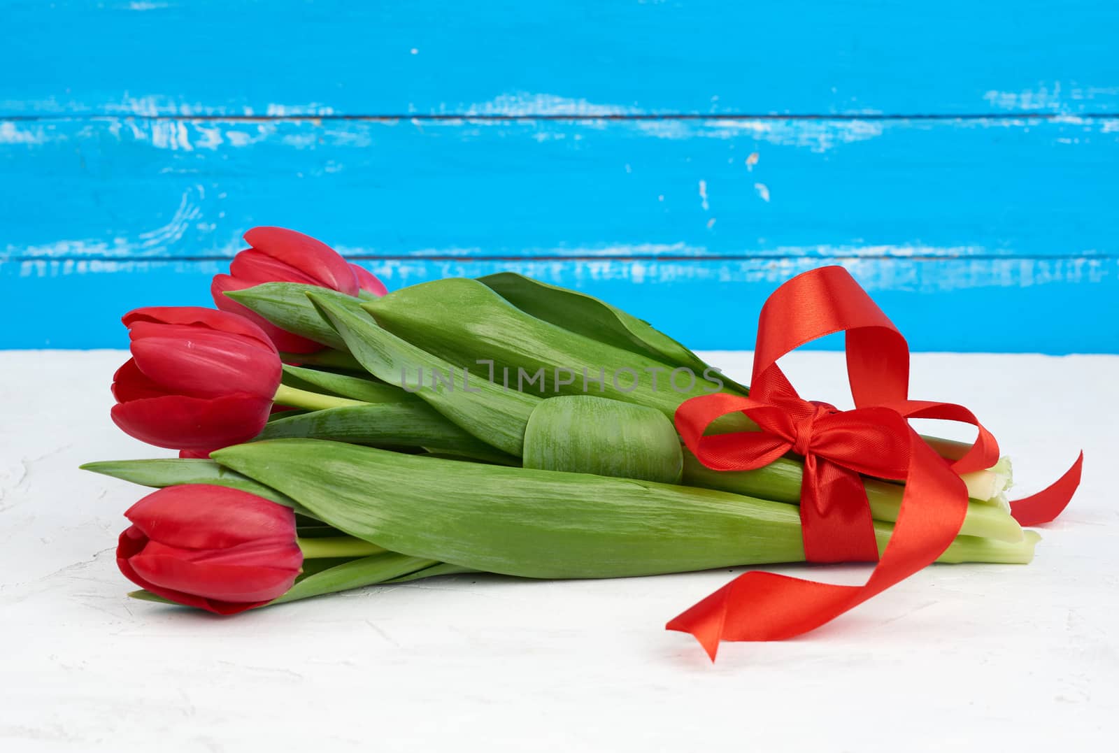 bouquet of red blooming tulips with green stems and leaves tied with a red silk ribbon, flowers lie on a blue wooden background, top view