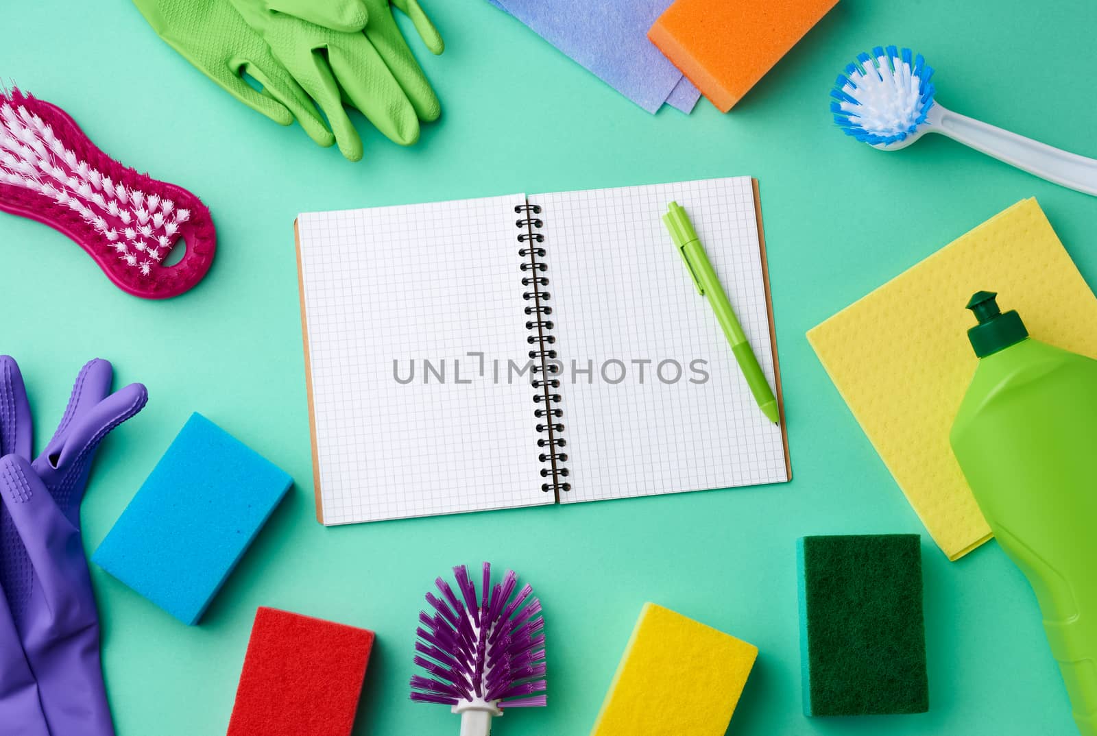 open notebook, rubber gloves for cleaning, multi-colored sponges, brushes and cleaning fluid in a green plastic bottle on a green background, place for text, flat lay