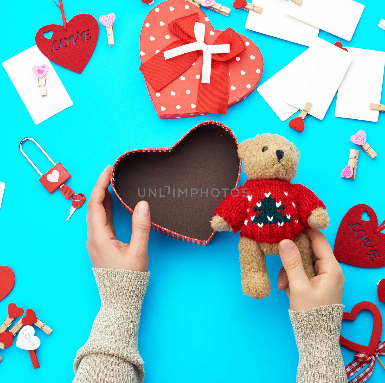 female hands holding a small brown teddy bear, on a blue background an open empty heart-shaped box, top view, holiday Valentine's Day