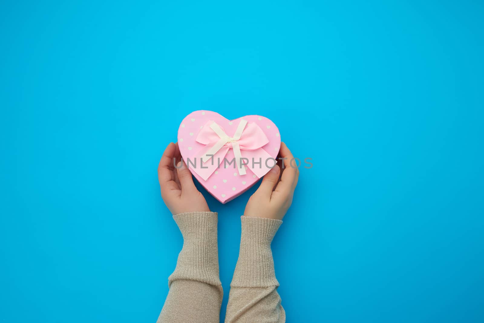 female hand holding a pink cardboard box with a bow on a blue background, concept of giving a gift for a holiday