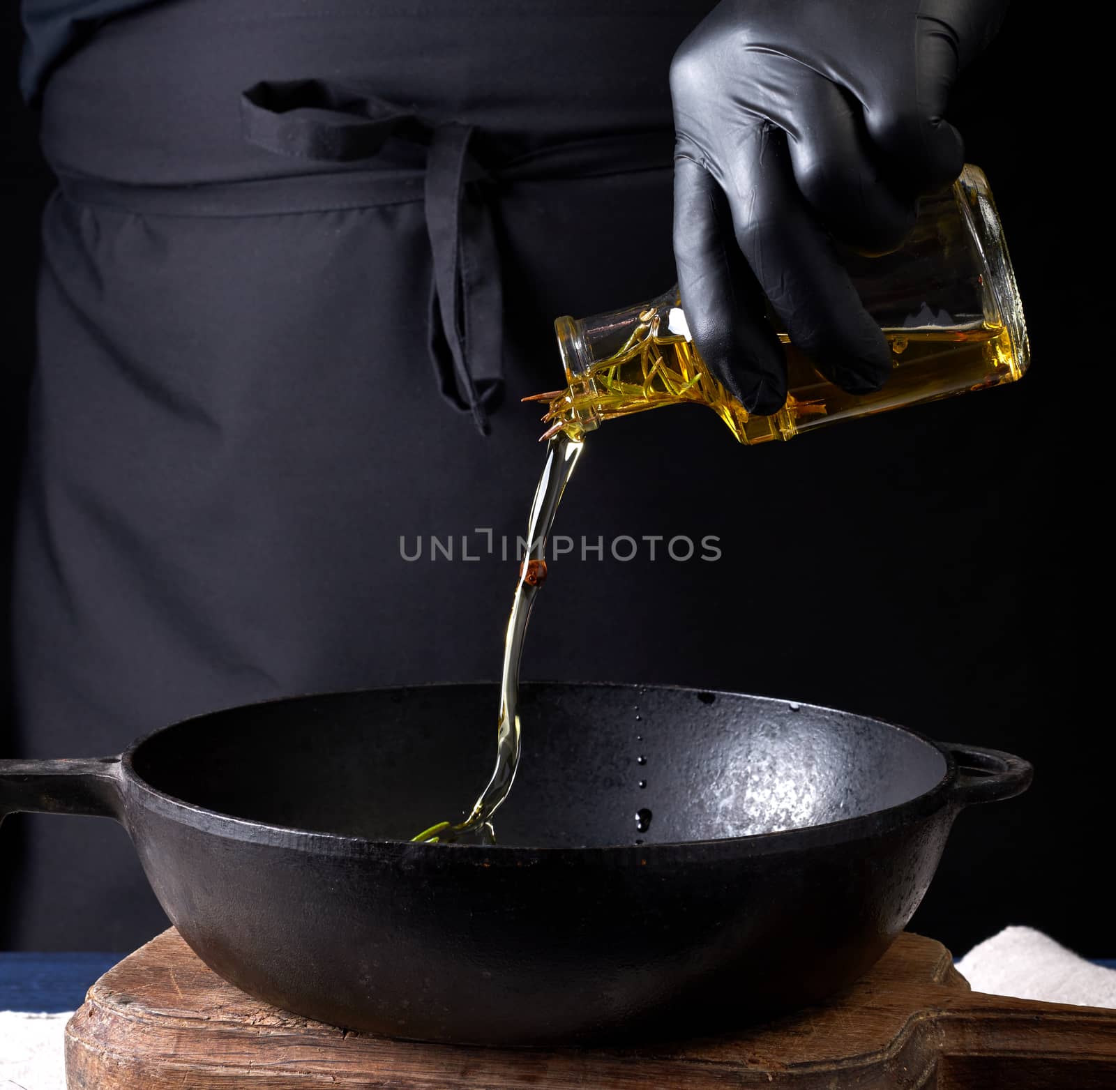 chef in black latex gloves pours olive oil from a transparent bottle into a black cast-iron frying pan, close up