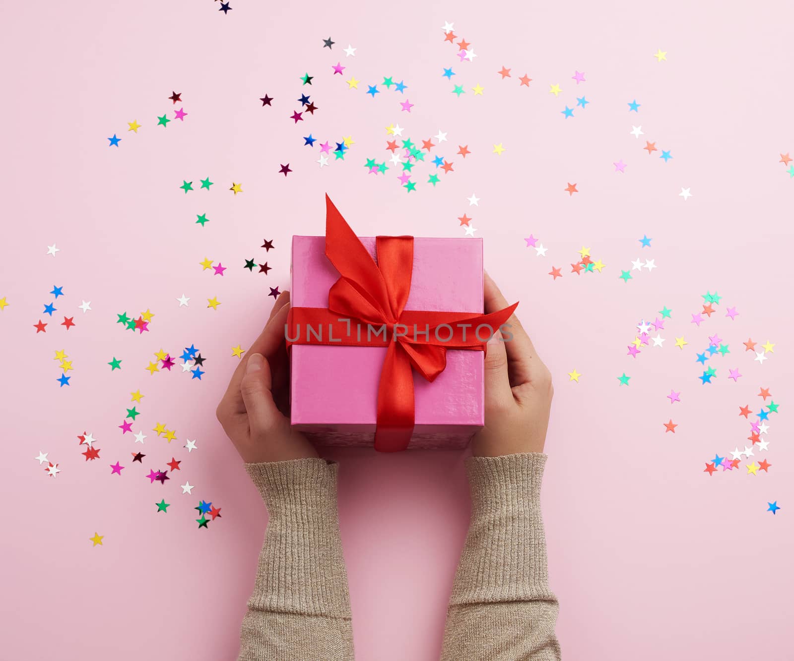 two female hands in a sweater hold closed square pink box with a bow on a pink background, top view, festive backdrop
