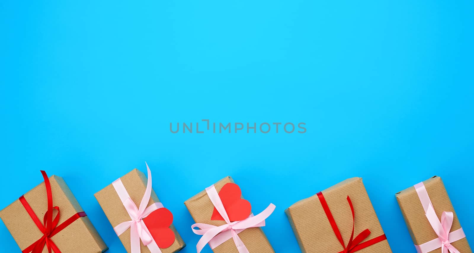 box is wrapped in brown paper and tied with a red thin silk ribbon on a blue background, red cut out paper hearts, copy space, holiday is Valentine's Day