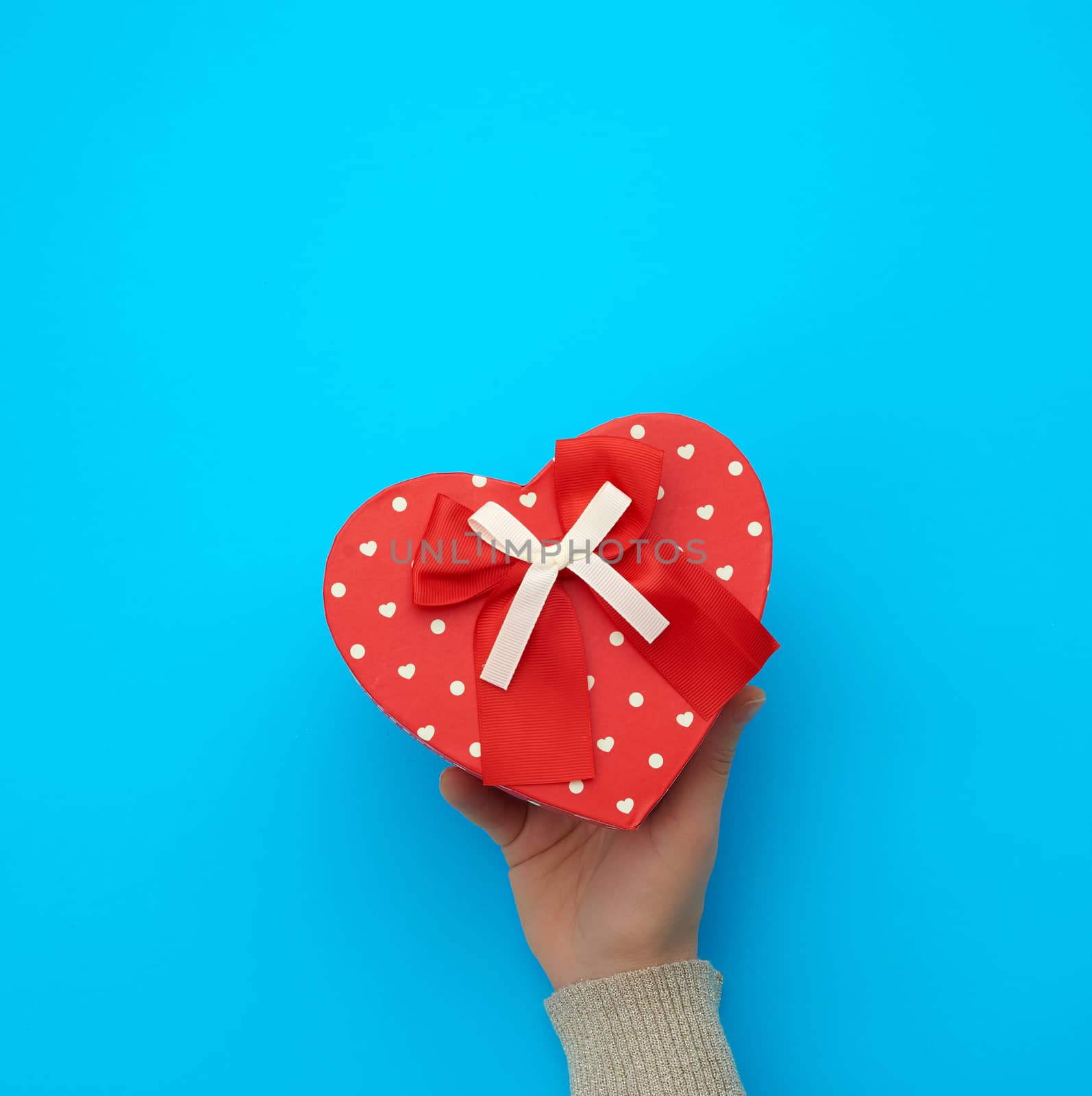 female hand holding a red cardboard box with a bow on a blue background, concept of giving a gift for a holiday