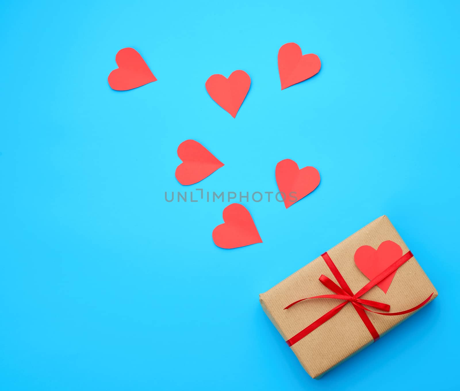 box is wrapped in brown paper and tied with a red thin silk ribbon on a blue background, red cut out paper hearts, copy space, holiday is Valentine's Day