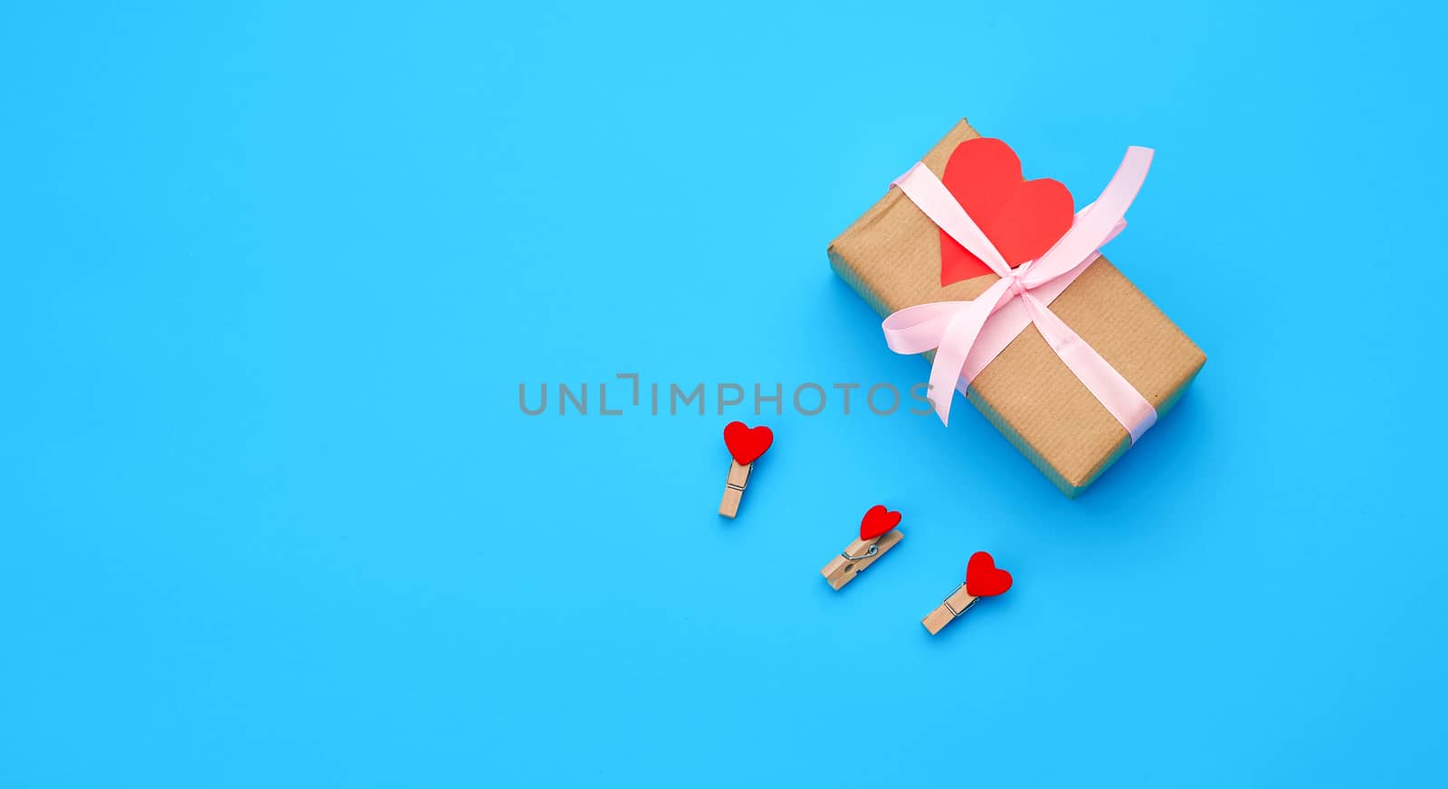 box is wrapped in brown paper and tied with a red thin silk ribbon on a blue background, red cut out paper hearts, copy space, holiday is Valentine's Day