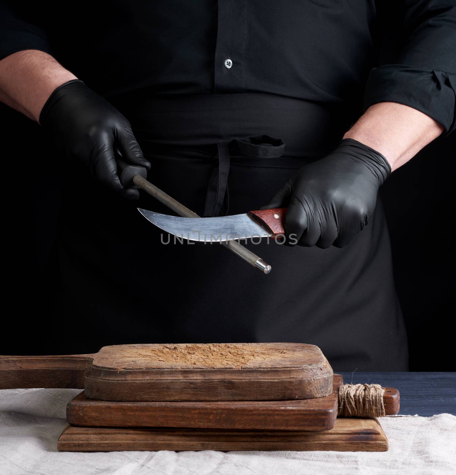 chef in a black shirt and black latex gloves sharpen a kitchen knife on an iron sharpener with a handle above the table, low key