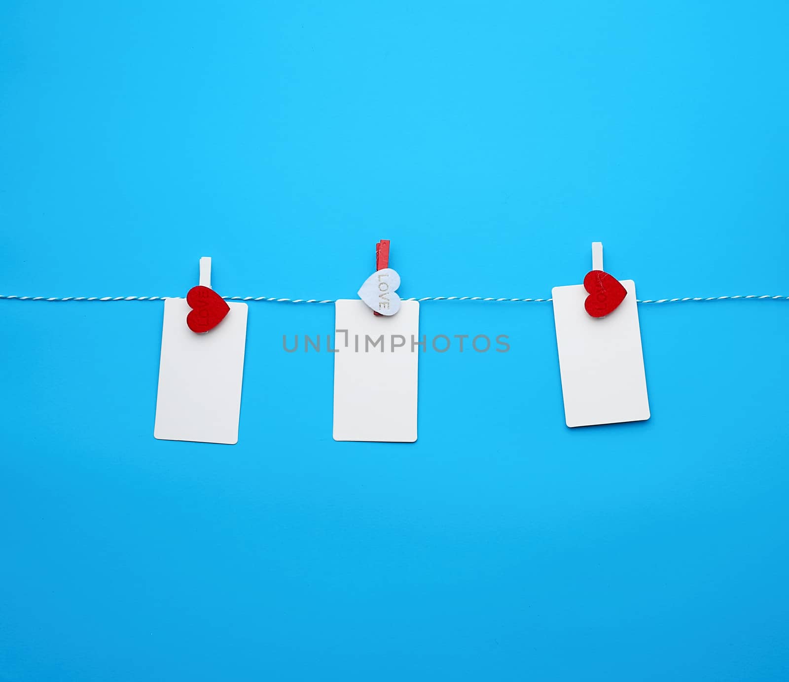 white empty paper rectangular business cards on decorative clothespins with a red heart hanging on a rope, blue background, place for an inscription