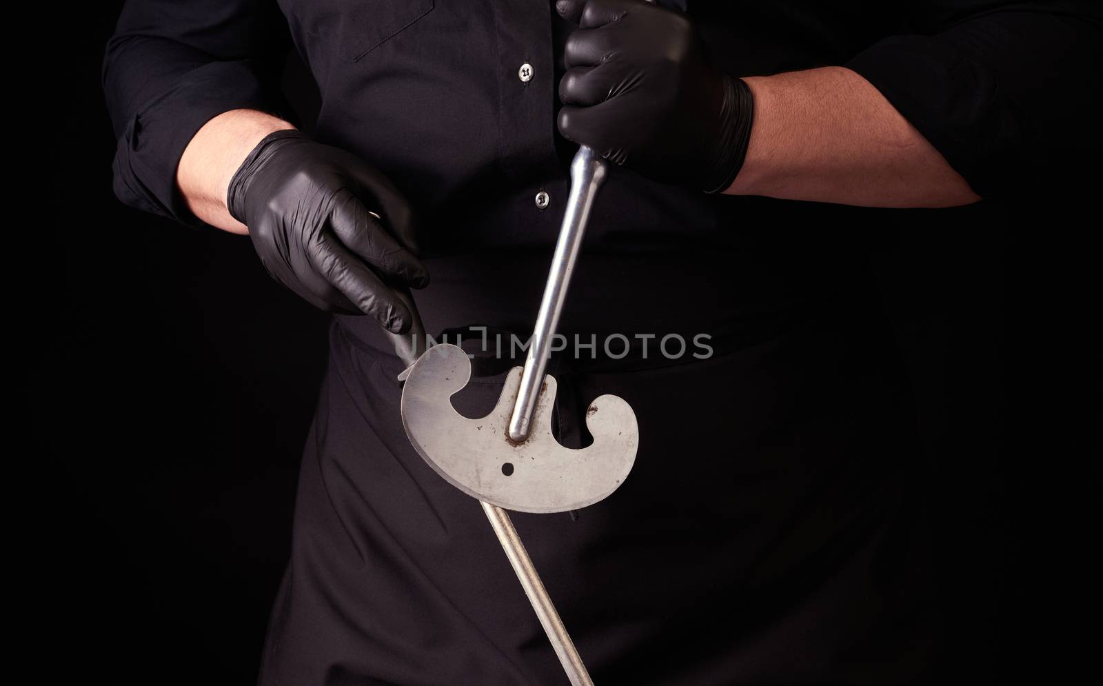 male cook in black uniform and black latex gloves holds a large sharp vintage knife for meat and vegetables and a metal sharpener for knives, black background