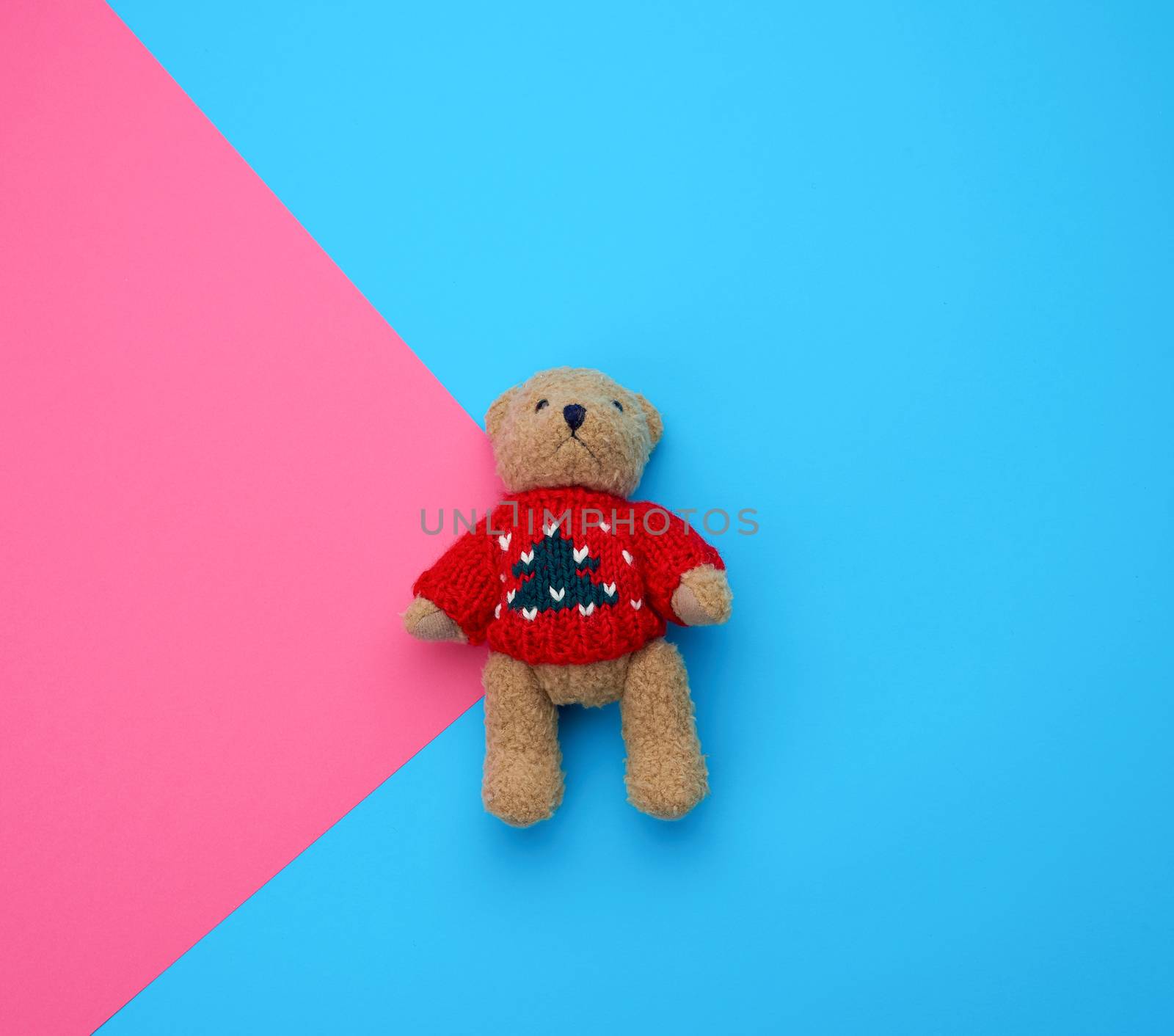 little cute brown teddy bear with in a red knitted sweater on a blue pink background, holiday toy