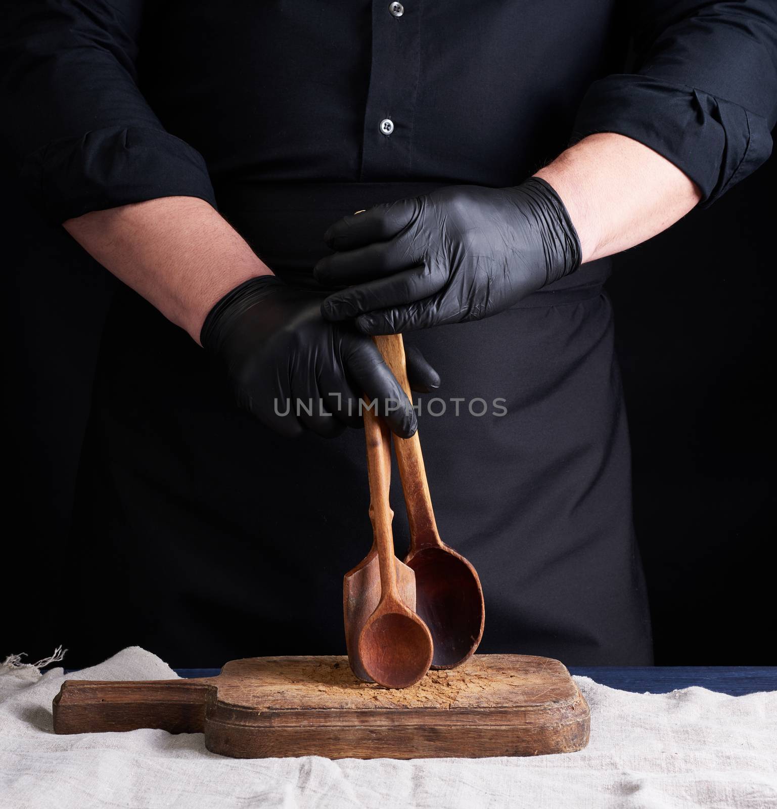chef in black latex gloves and a black uniform holds wooden vintage spoons in front of him, low key