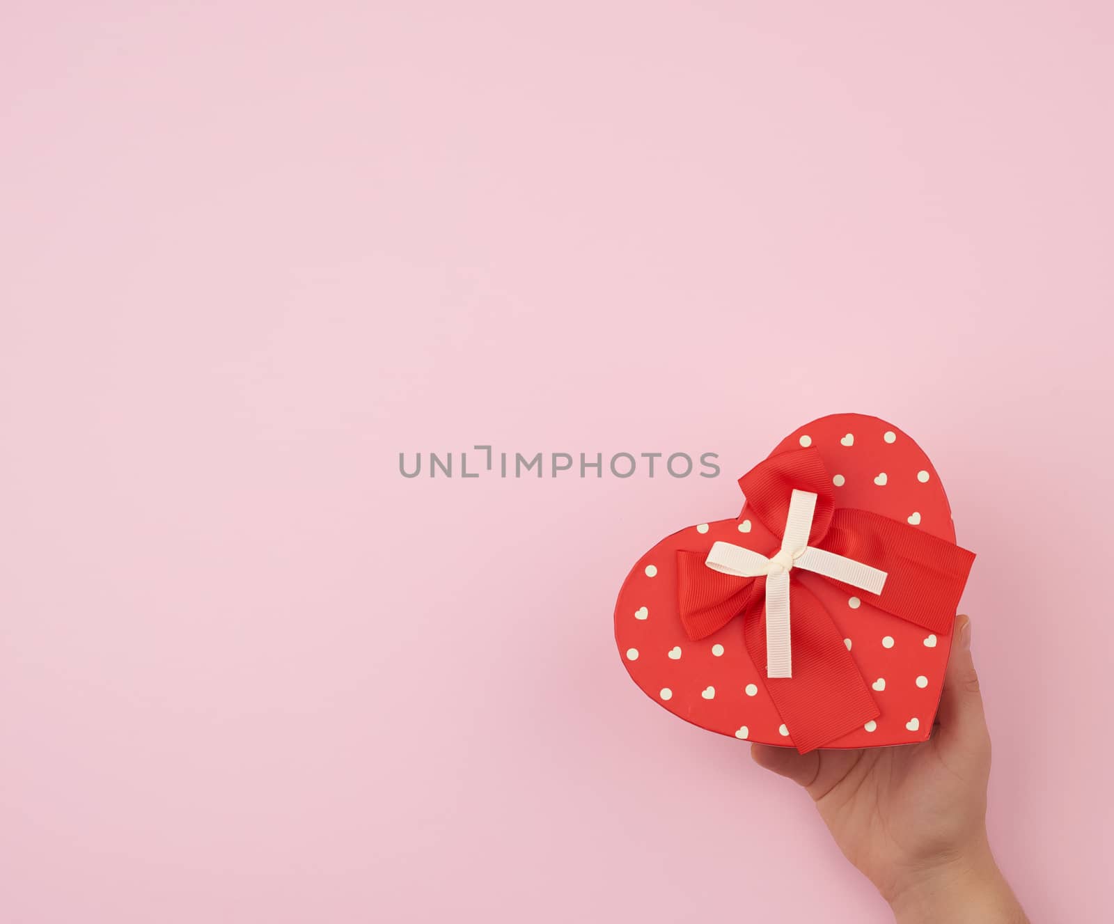 female hand holding a red cardboard box with a bow on a pink background, concept of giving a gift for a holiday