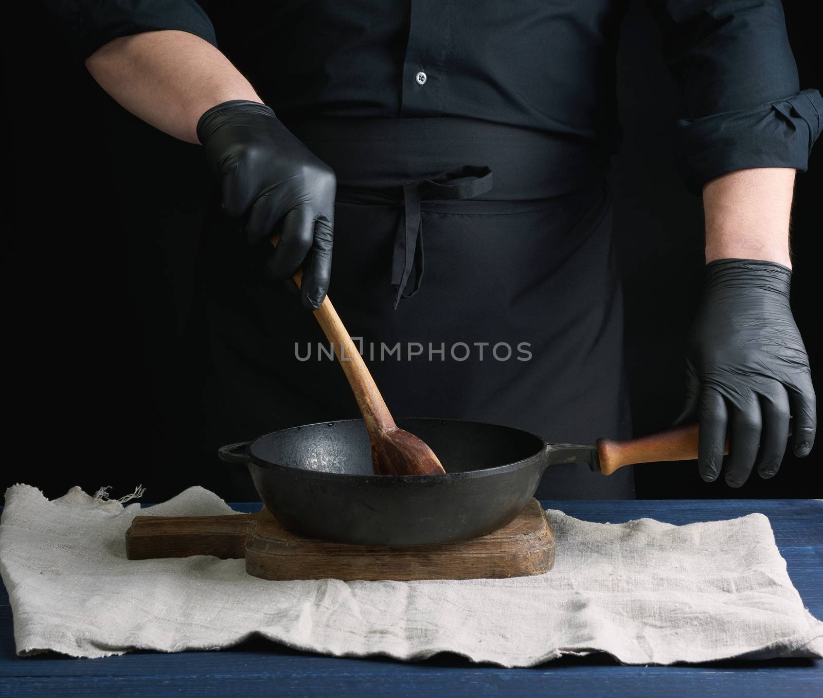 male cook in black uniform and latex gloves stirs a vintage wooden spoon food in a cast-iron pan with a handle, dark background