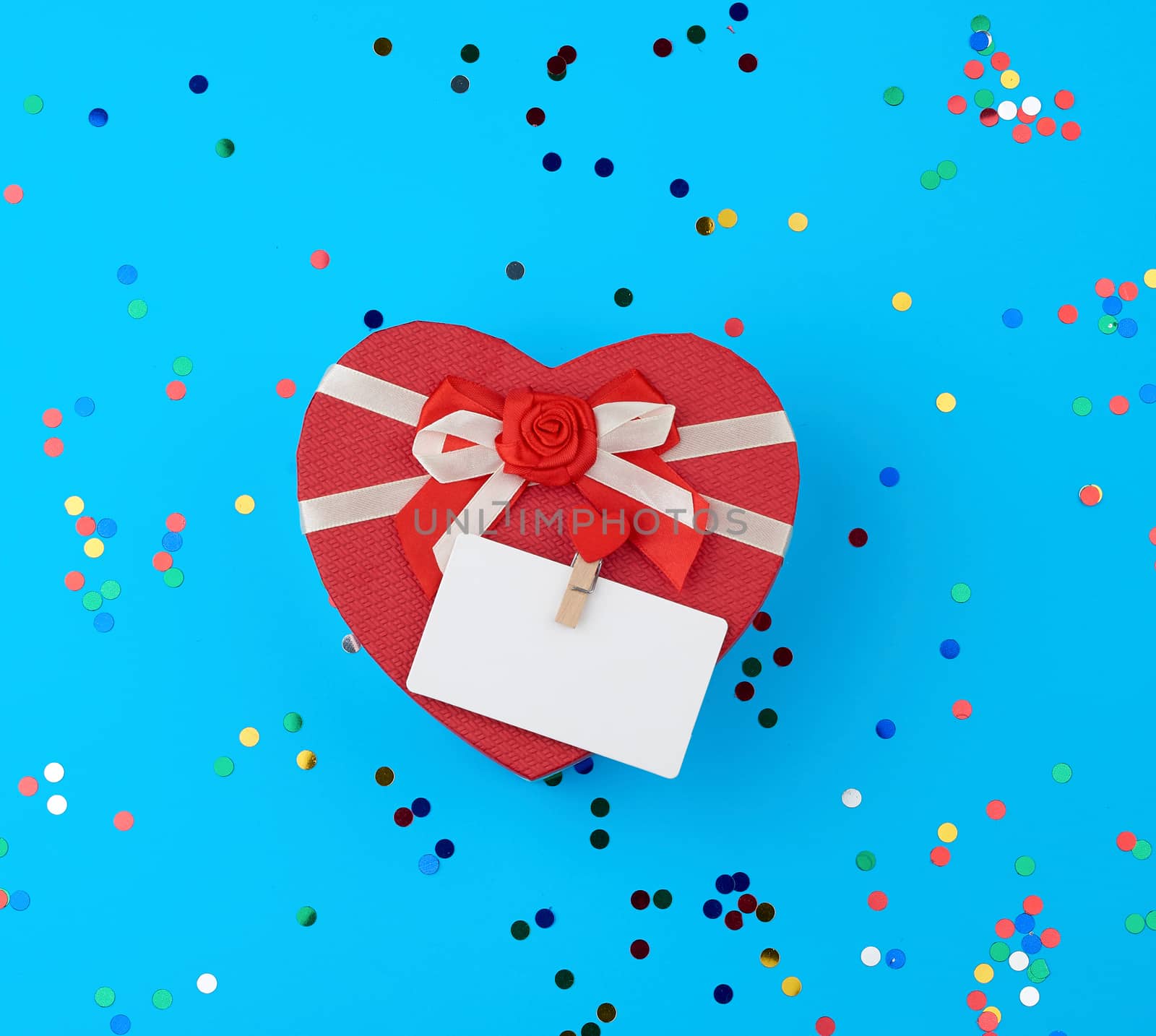closed red heart-shaped cardboard box on a blue background with multi-colored shiny confetti, festive backdrop for birthday, Valentine's day