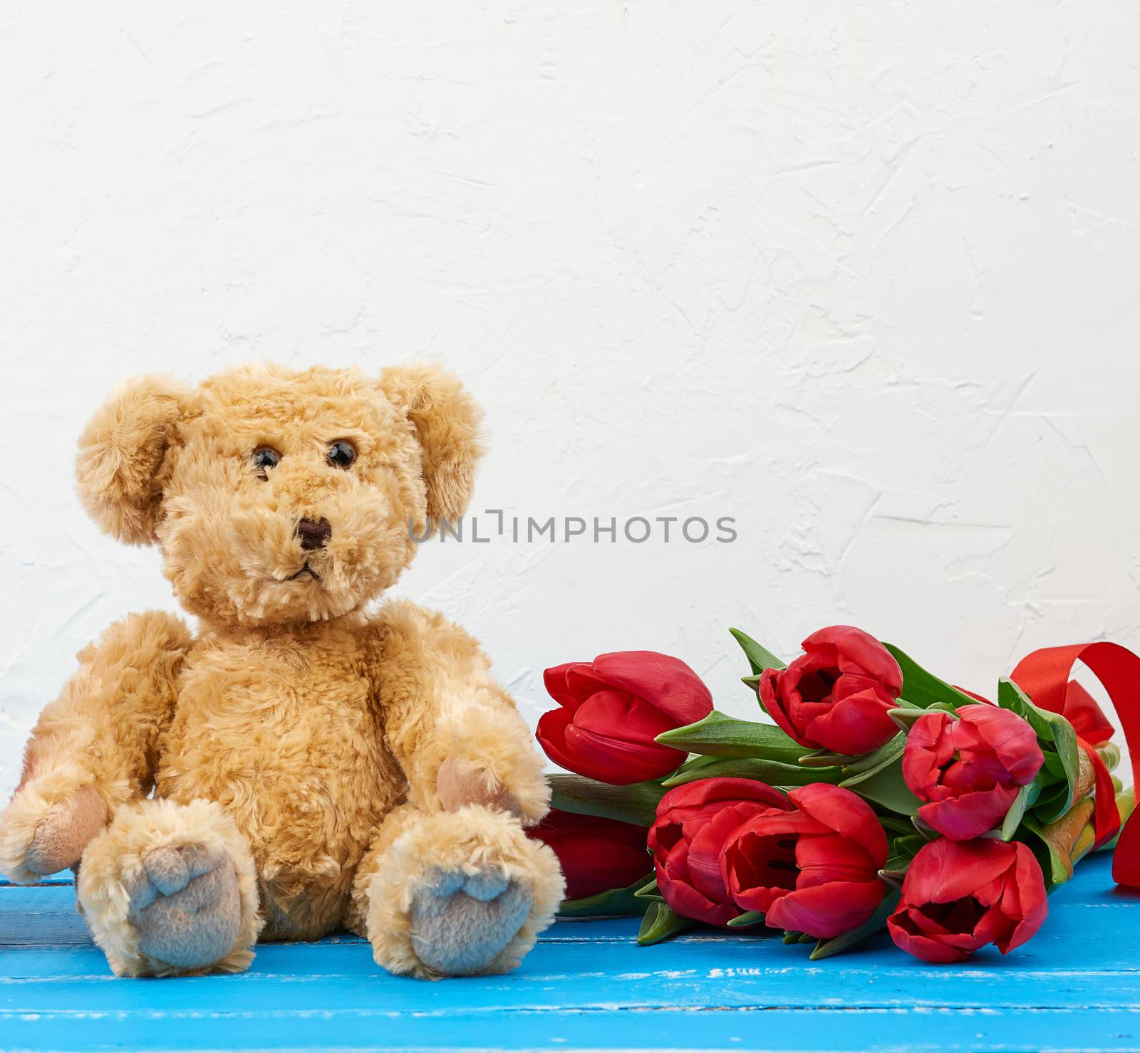 cute brown teddy bear sits on a blue wooden background and bouquet of red tulips, festive backdrop for birthday, Valentine's day, anniversary
