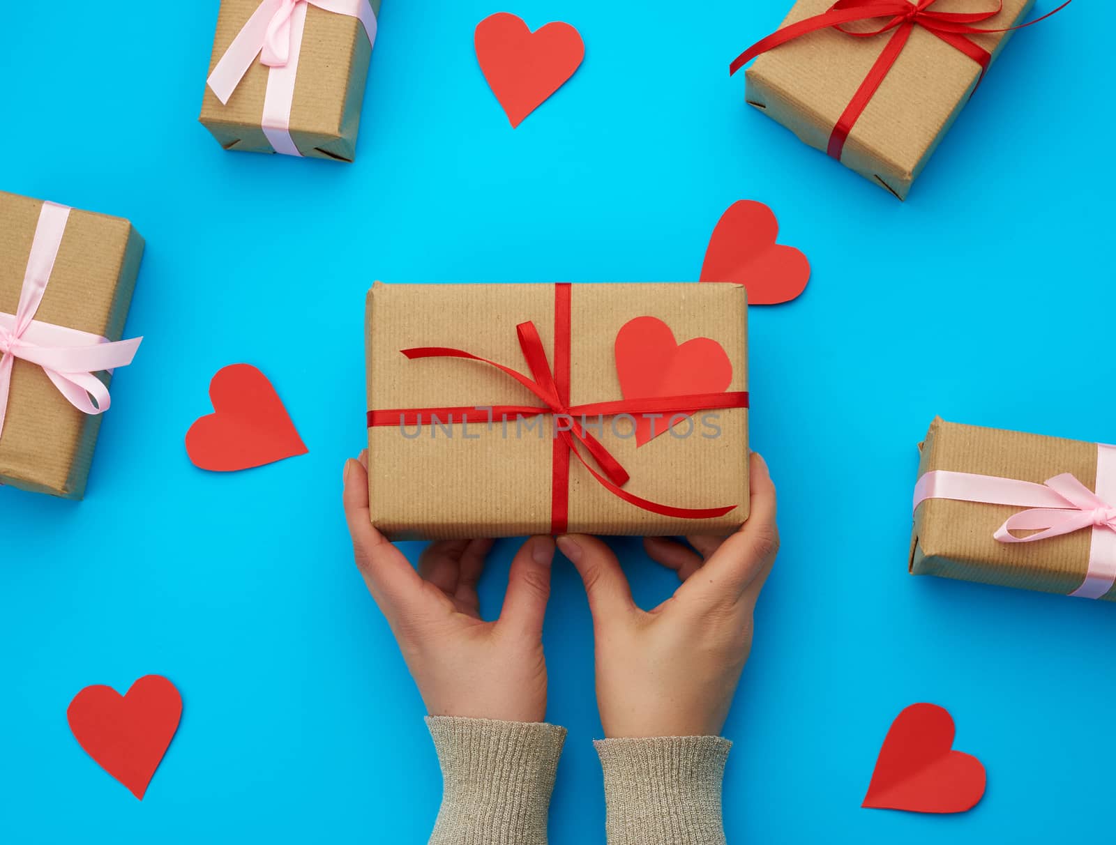 female hand holds a box wrapped in brown kraft paper and tied with a silk ribbon on a blue background, paper red hearts. Valentine's Day backdrop
