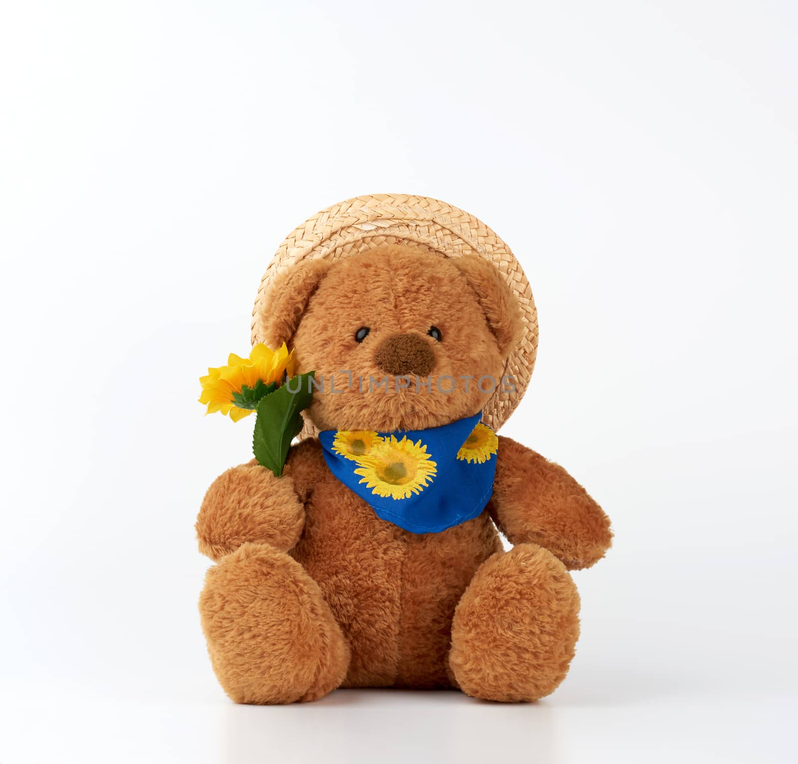 cute little brown teddy bear holds a plastic yellow sunflower and is dressed in a straw hat,  toy is sitting on a white background