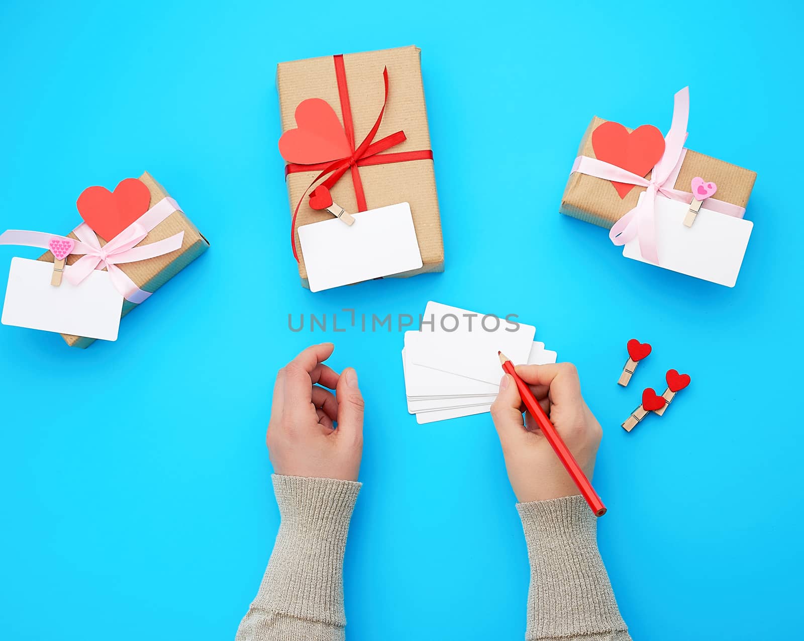 female hand holds a box wrapped in brown kraft paper and tied with a silk red ribbon on a blue background, paper red hearts. Valentine's Day backdrop