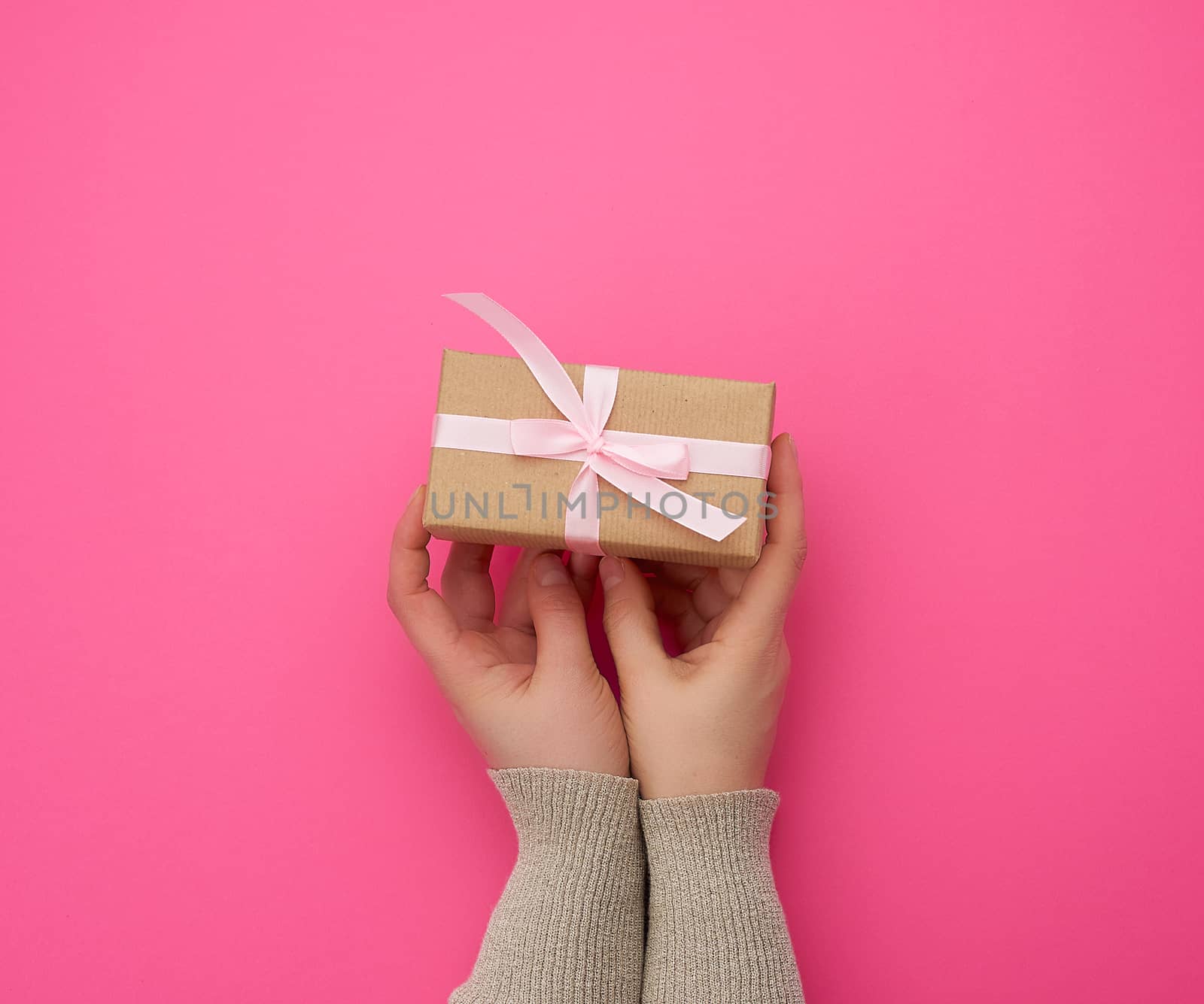 female hand holds a brown box with a red bow on a pink background, concept of a congratulation on a happy birthday, place for text