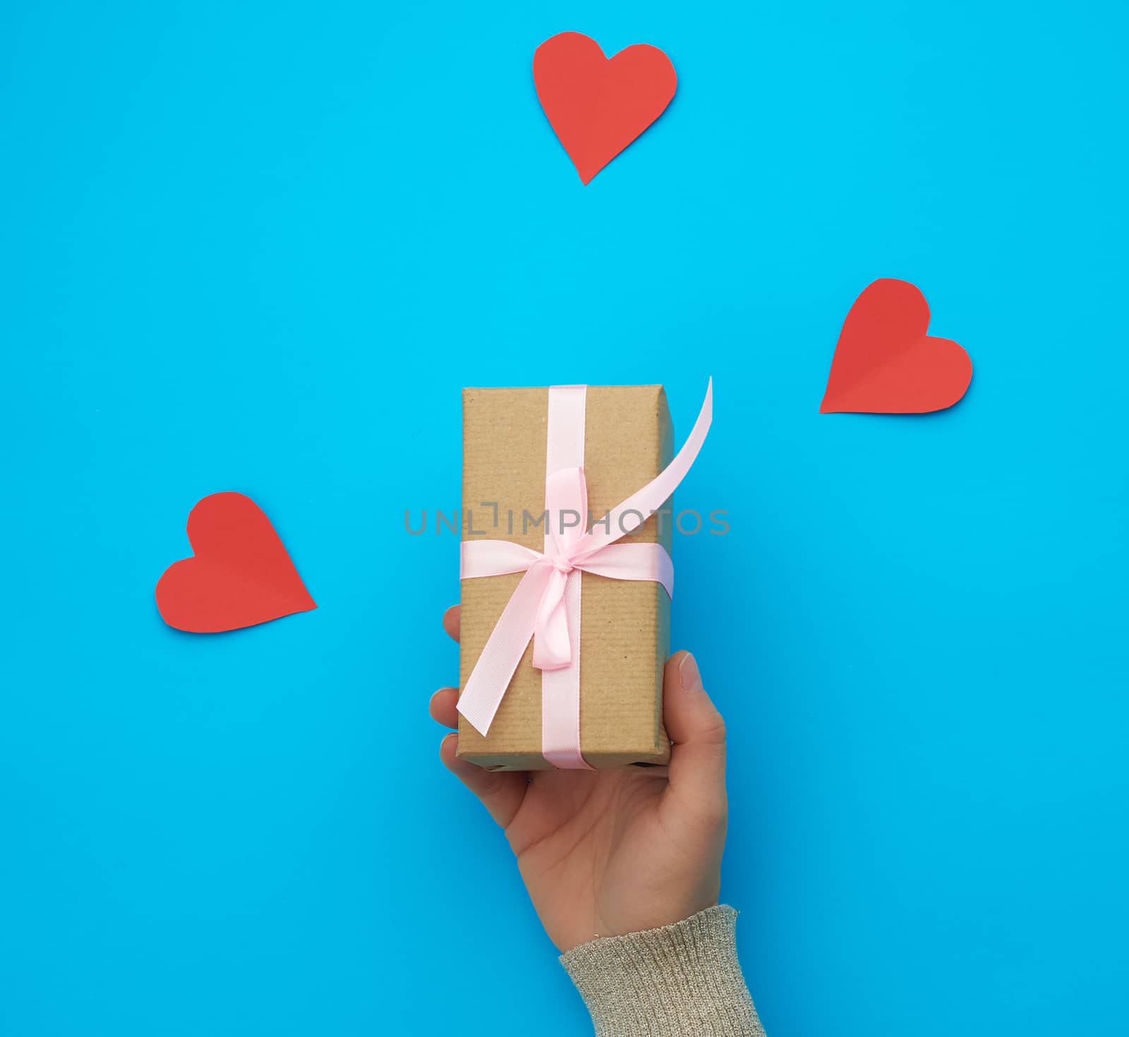 female hand holds a box wrapped in brown kraft paper and tied with a silk ribbon on a blue background, paper red hearts. Valentine's Day backdrop