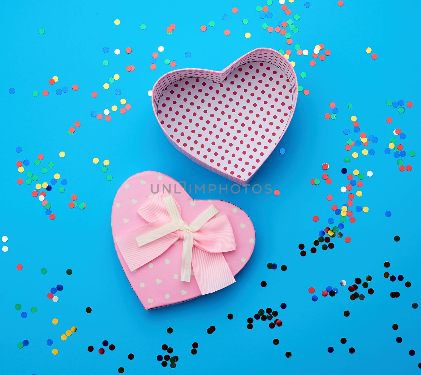 open pink heart-shaped cardboard box on a blue background with multi-colored shiny confetti, festive backdrop for birthday, Valentine's day