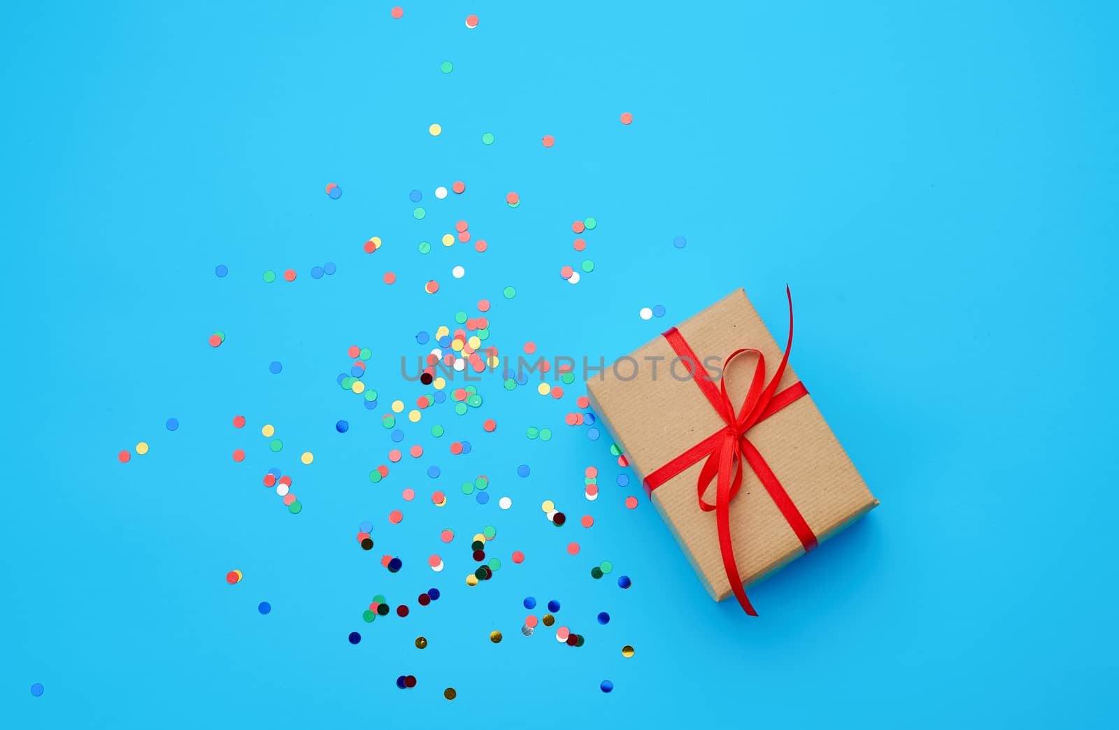 brown gift box wrapped in paper and tied with silk ribbon on a blue background with multi-colored shiny confetti, festive backdrop for birthday, valentine's day