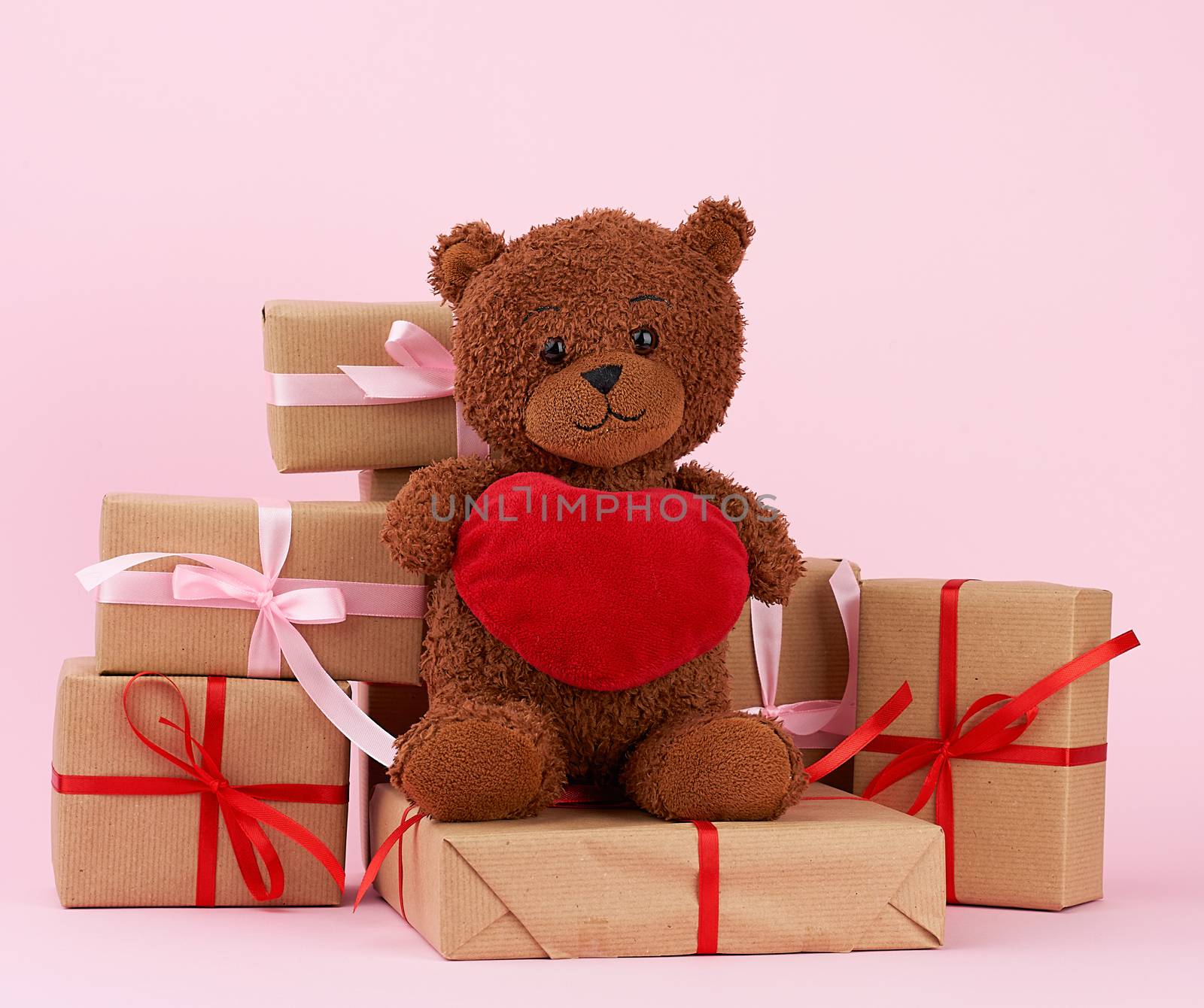 cute brown teddy bear and gift in box wrapped in brown eco paper, tied with silk red ribbon on a pink background, happy birthday and valentines day