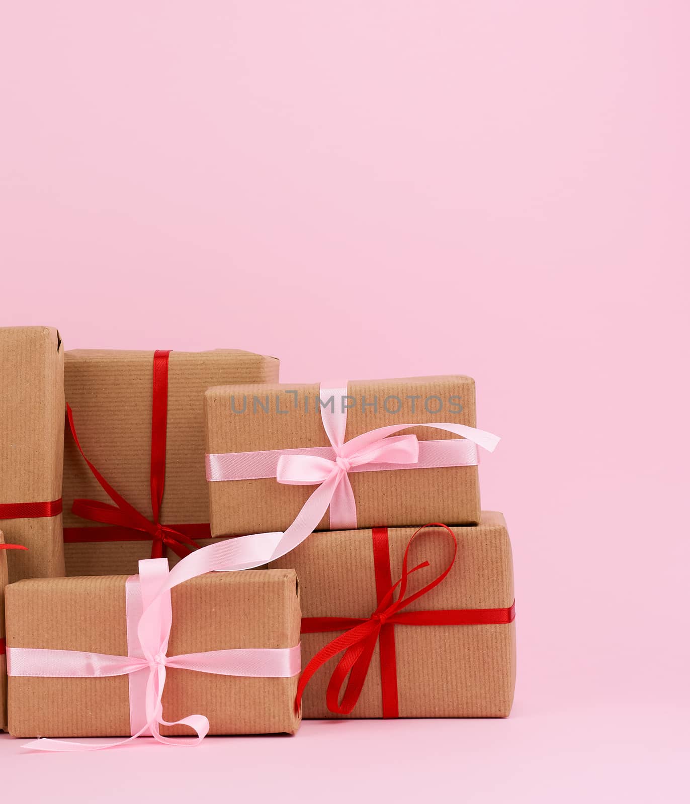 stack of gifts in boxes wrapped in brown kraft paper and tied with silk ribbon on a pink background. Festive concept, copy space