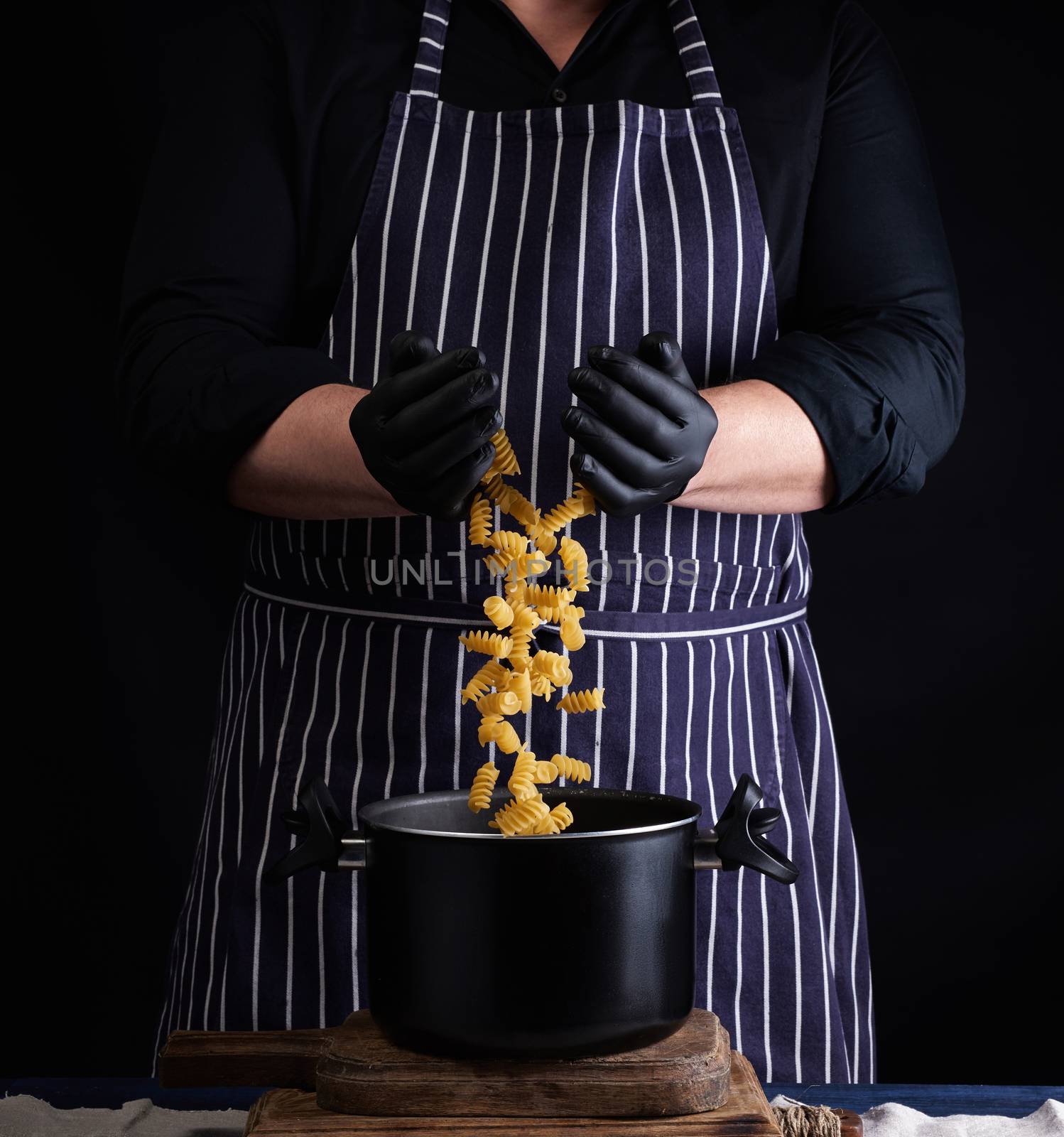 chef in black latex gloves, striped apron pours raw fusilli pasta into a metal pan, cooking process, twisted pasta frozen in the air